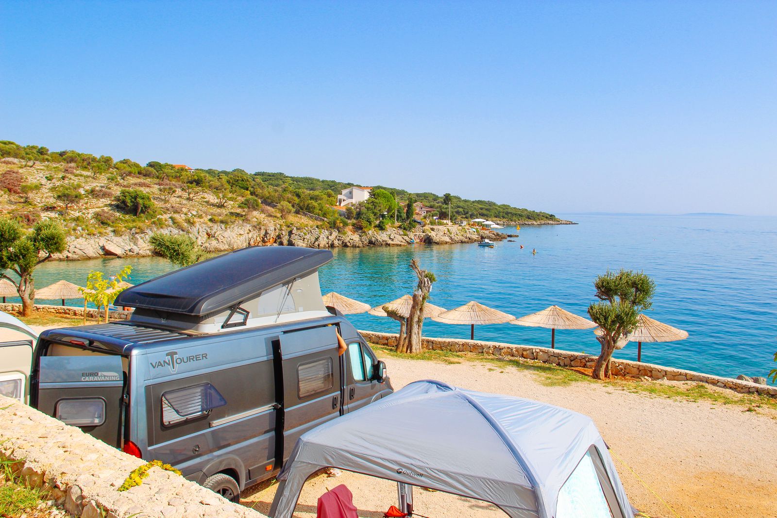 Wohnmobil mit Aufstelldach an einer Bucht der kroatischen Adriaküste mit Sonnenschirmen und türkisfarbenem Wasser